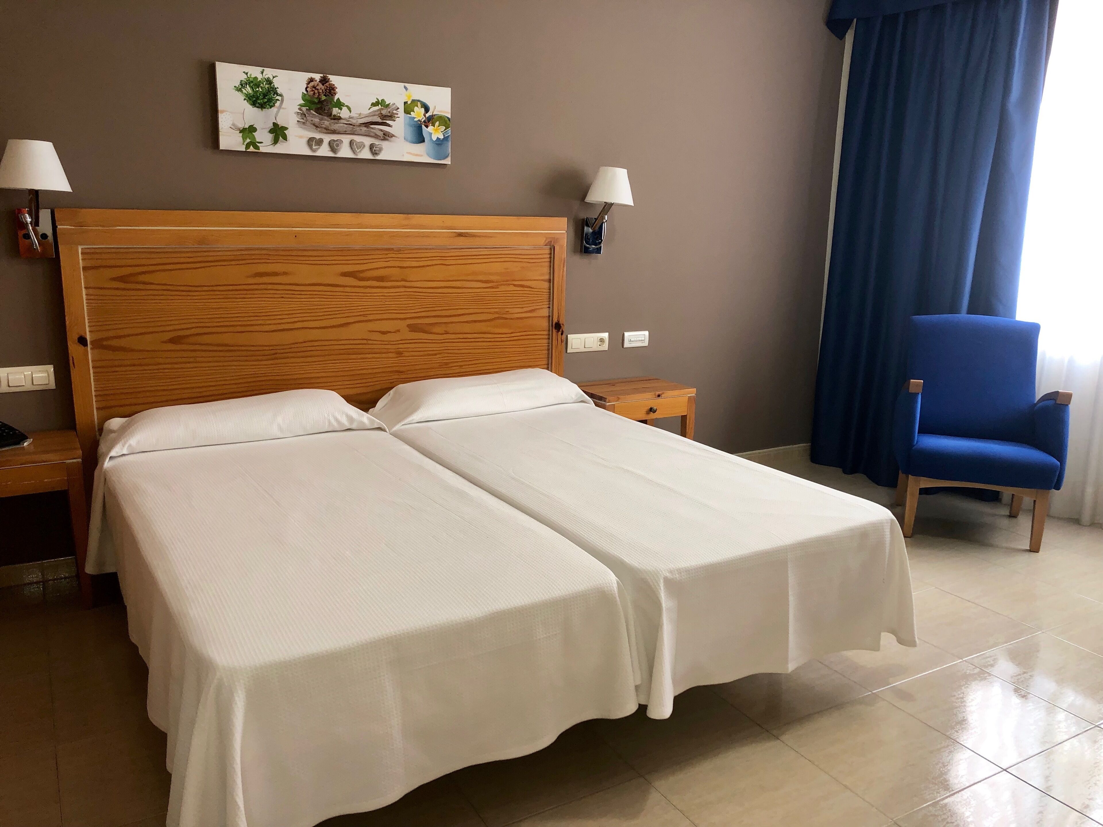 Habitación de hotel con una cama doble, dos mesitas de noche, un sillón azul, una lámpara de pared y un adorno en la pared.