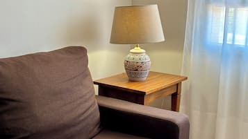 Un canapé marron, une table d’appoint en bois surmontée d’une lampe à motifs et une fenêtre ornée de rideaux légers.