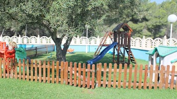 Une aire de jeux avec un toboggan et une structure d’escalade, entourée d’une clôture en bois et d’arbres.