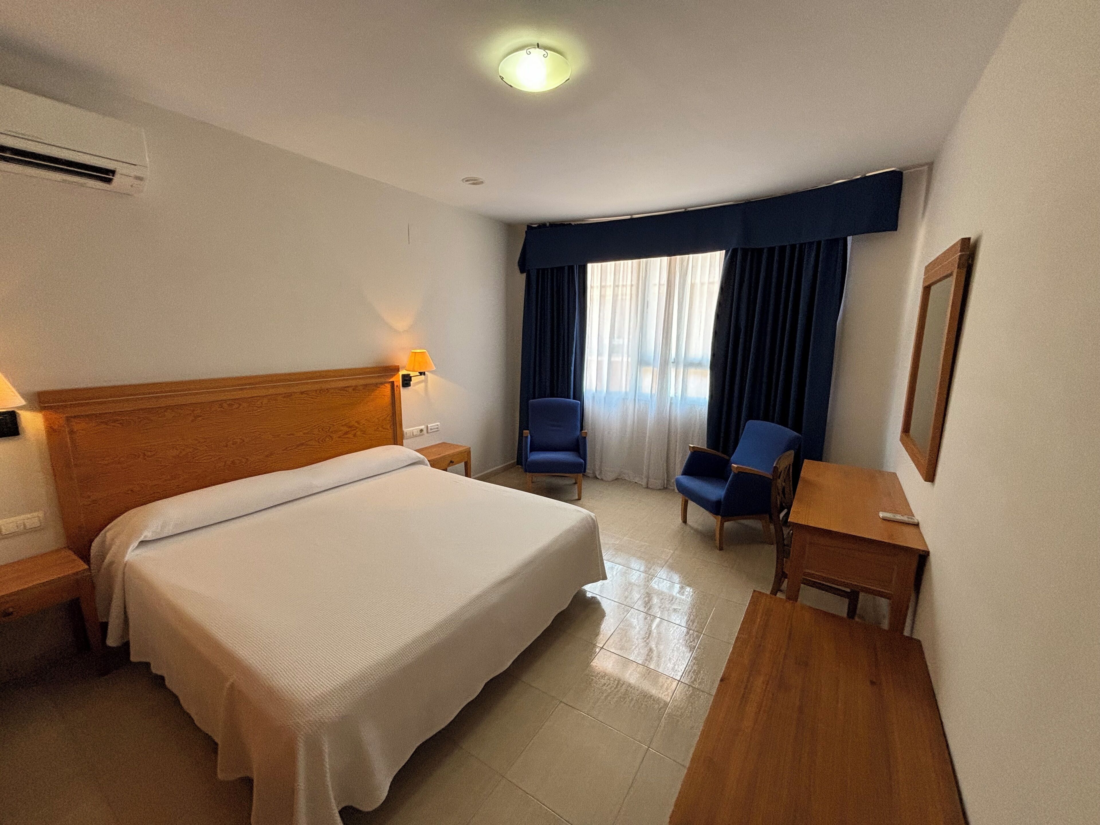 A hotel room with a large bed, a desk, two chairs, a lamp, and a window with curtains.