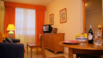 A living room with a sofa, a television, a wooden cabinet, a dining table with a bowl of fruit, and a wine bottle.