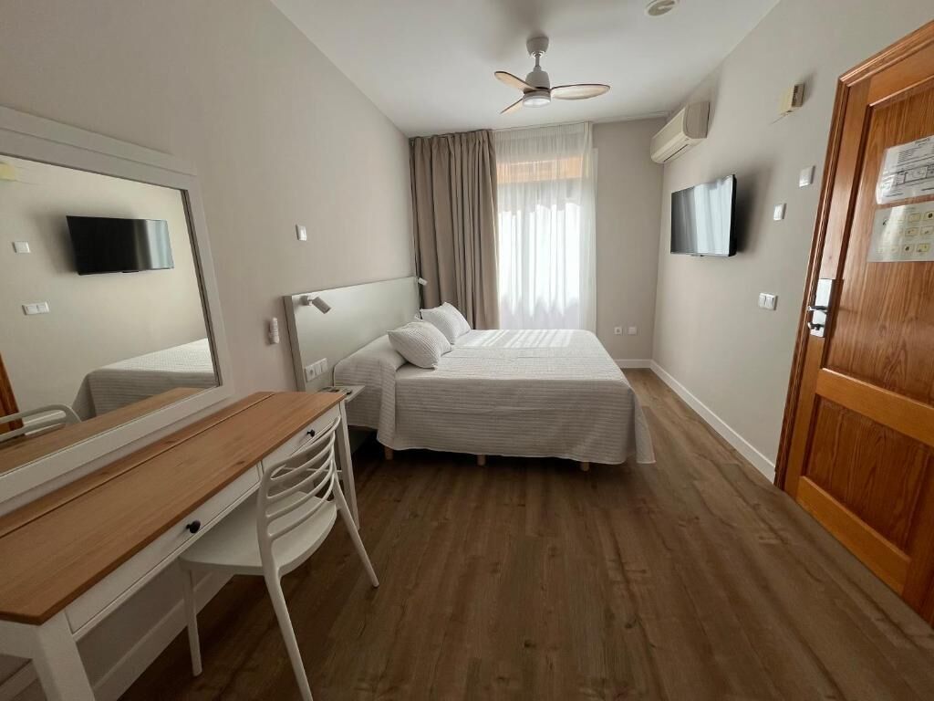 A hotel room with a bed, a desk, a chair, a ceiling fan, a TV, and a wooden door.