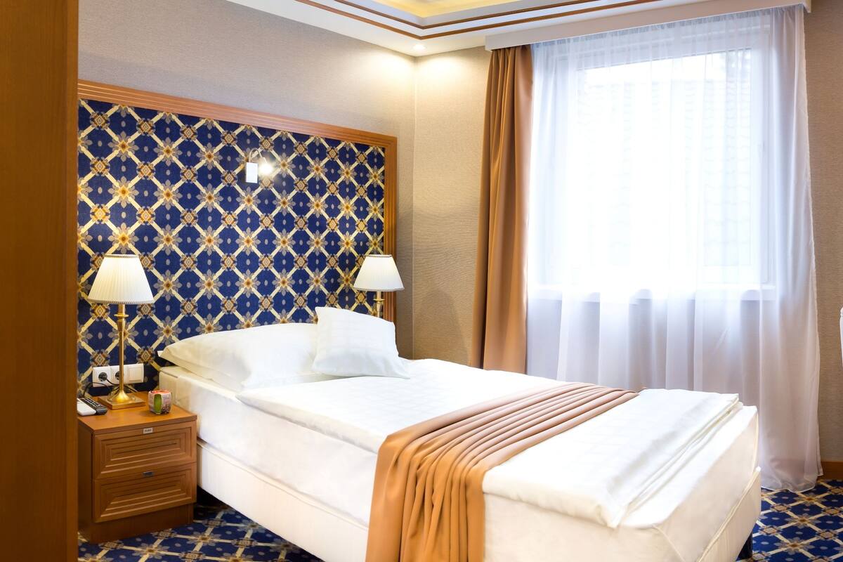 Hotelkamer met een bed, nachtlampjes, een bureau met telefoon en koffiezetapparaat, en een groot raam met gordijnen.