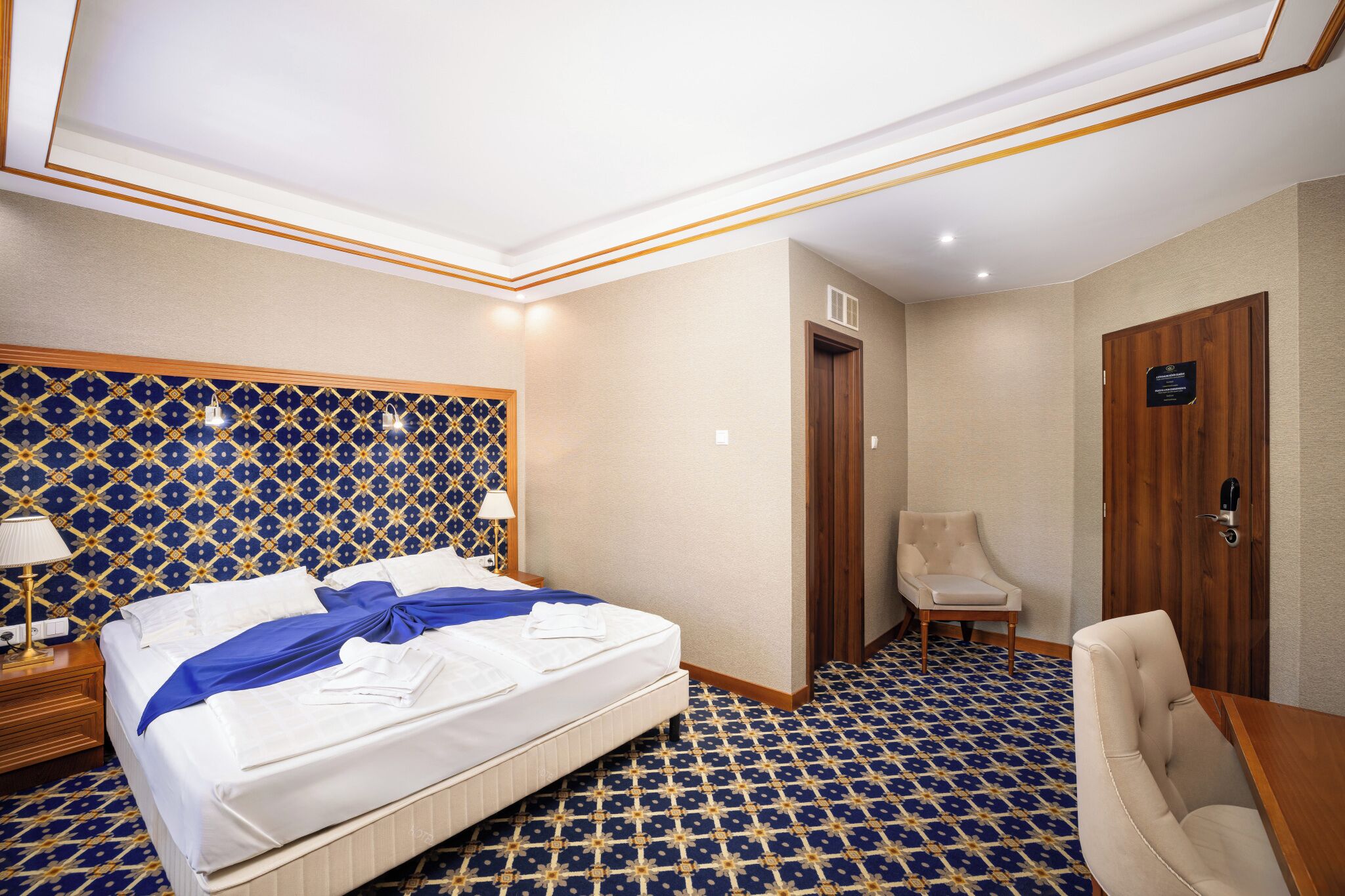 A hotel room with a patterned floor, a bed with white linens, a wooden headboard, a wooden door, and a chair.
