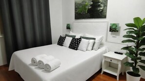 Cama bem arrumada com roupa de cama branca e almofadas pretas e brancas de bolinhas, uma pequena mesa de cabeceira com um controle remoto e uma planta em vaso à direita.