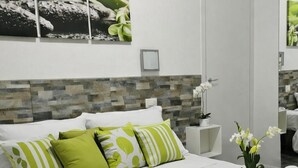 Quarto de hotel moderno com uma cama bem arrumada, uma pequena mesa de cabeceira com um vaso de flores e um espelho grande.