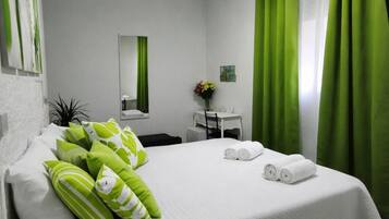 Ein Hotelzimmer mit einem Bett, grünen Kissen, einem Schreibtisch, einem Stuhl, einem kleinen Tisch mit Blumenvase und grünen Vorhängen.