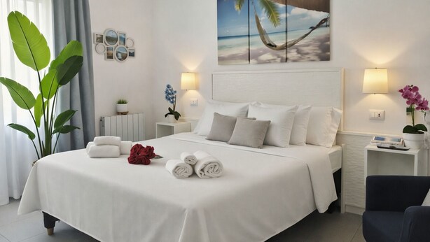 Quarto de hotel com uma cama grande, mesa de cabeceira com abajur, quadro na parede retratando uma praia e um vaso com planta.