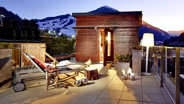 Een houten chalet met een balkon, voorzien van een ligbed, een tafel met kaarsen, en uitzicht op besneeuwde bergen.