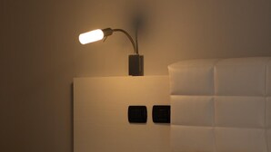 Una cama bien tendida con edredón blanco, manta negra y una toalla blanca doblada. Se observa una lámpara de pared con brazo curvo y base rectangular.