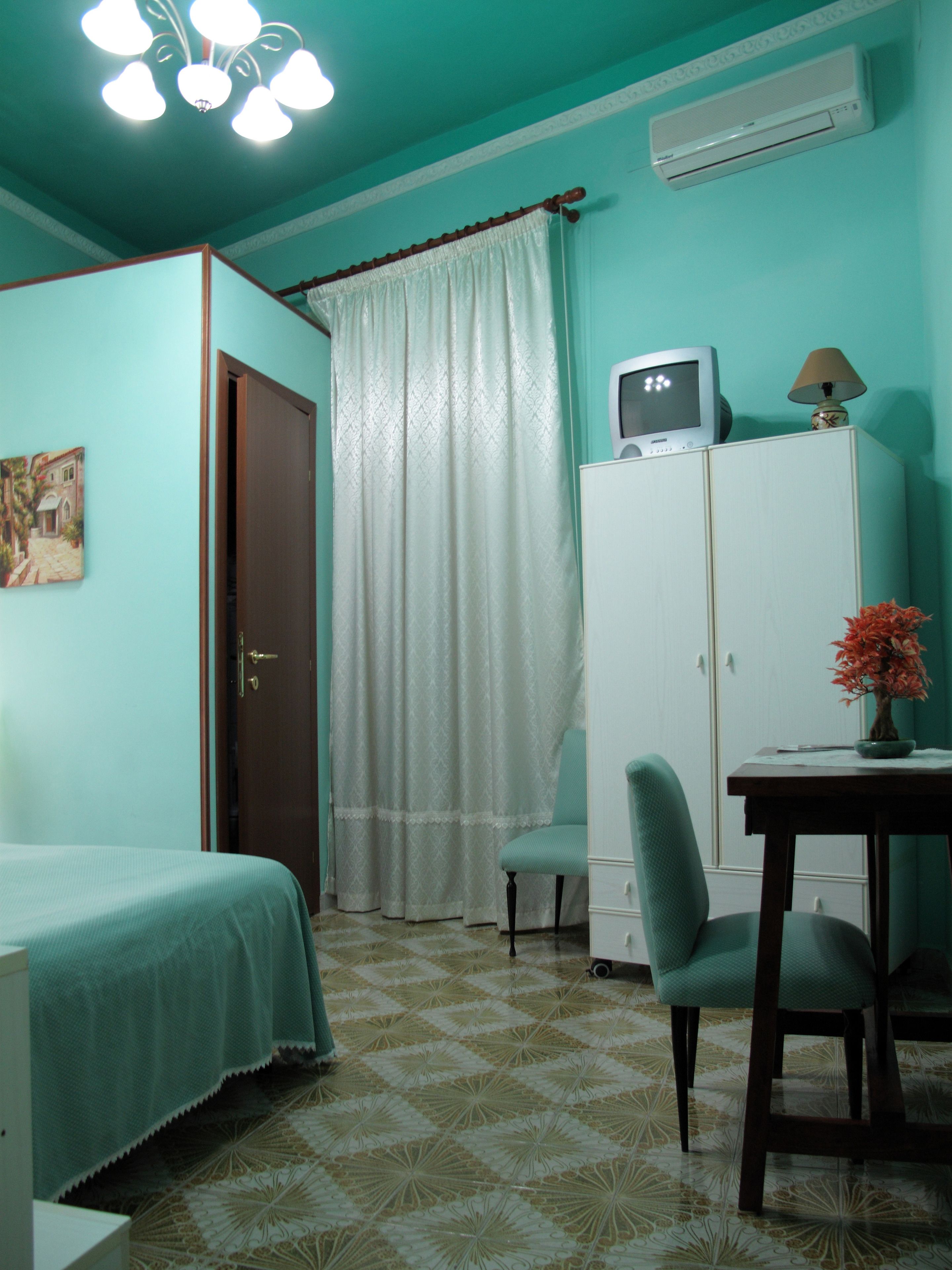 A room with teal walls, a bed, a desk with a lamp, a chair, and a television on a cabinet.