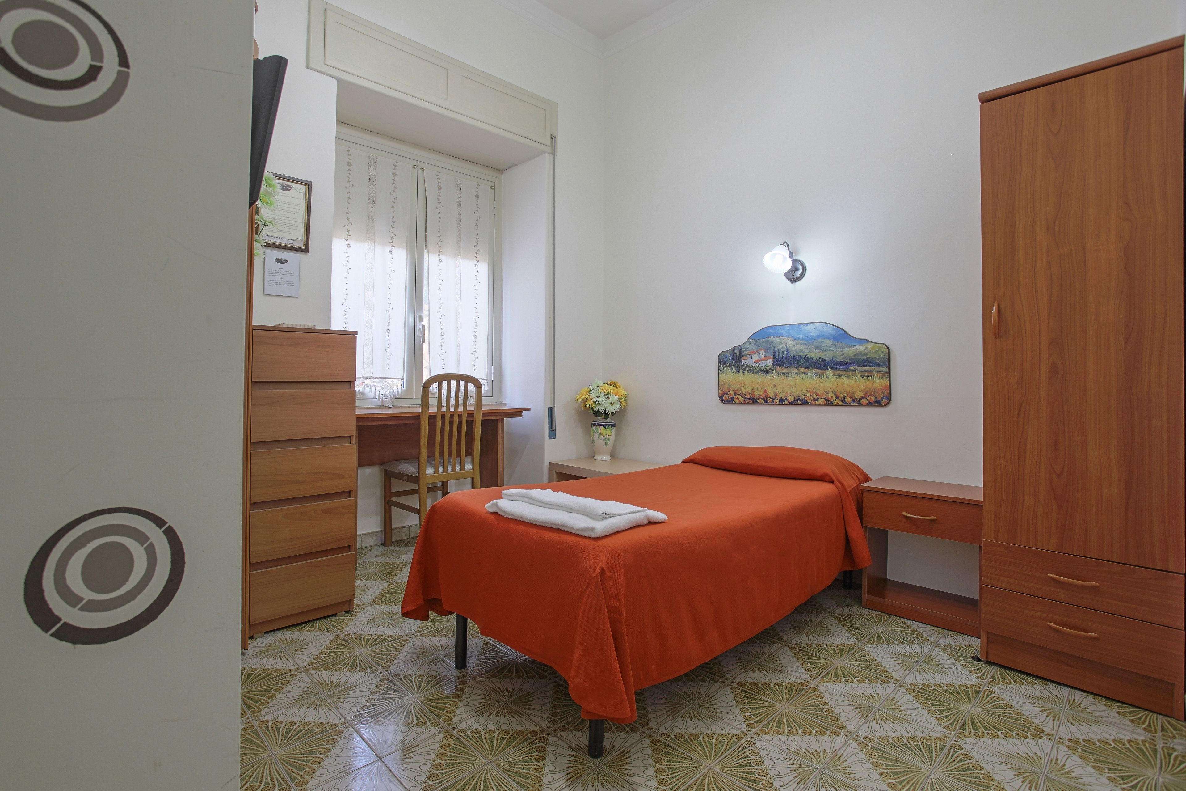 A bedroom with a bed, a wooden wardrobe, a desk, a chair, and a small table with a vase of flowers.