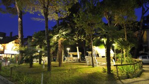 An outdoor seating area with tables and chairs, surrounded by trees and illuminated by ambient lighting. - Mimosa (Lignano Sabbiadoro)