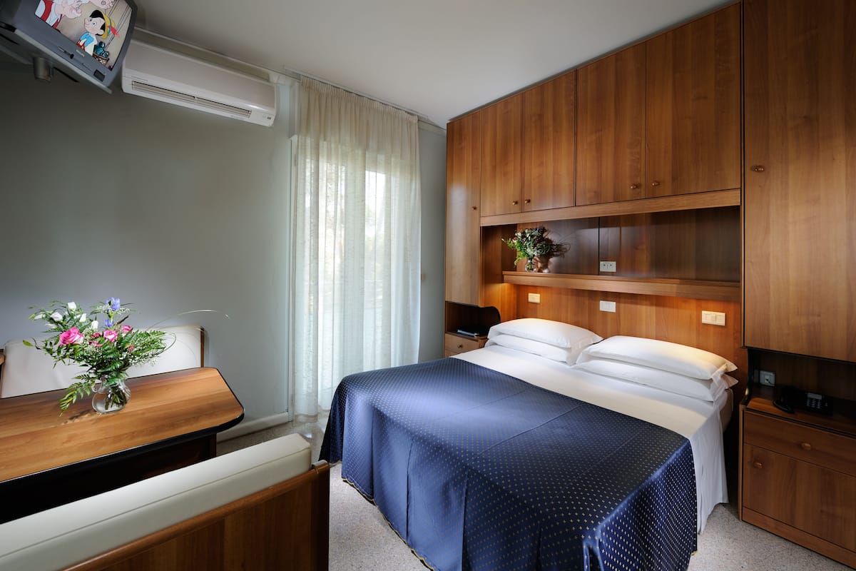 A hotel room with a bed, bedside table, and a small desk with a vase of flowers.