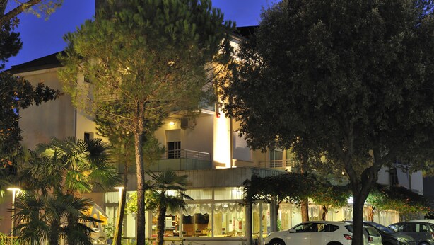 A hotel with a two-story structure, a covered outdoor seating area, and a parking lot with several cars. - Mimosa (Lignano Sabbiadoro)