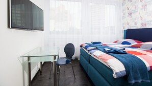 A modern bedroom with a glass desk, a bed with a colorful quilt, and a cityscape view through sheer curtains. - Design Sleepy Cologne (Cologne)