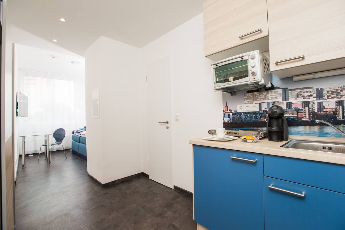 Cocina moderna con microondas, cafetera y lavabo. Se aprecia un armario azul y una ventana con vistas a la ciudad.