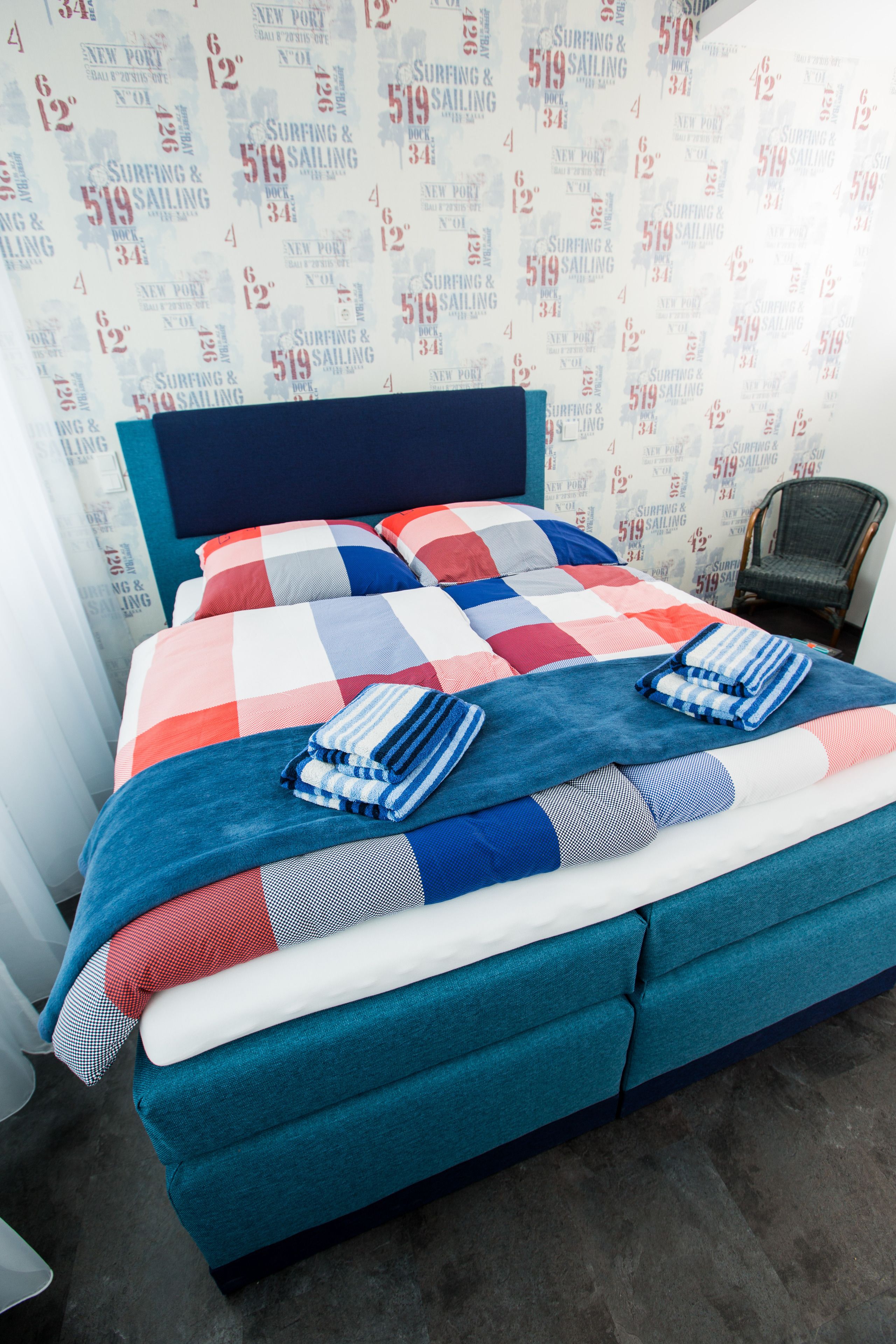 A neatly made bed with a checkered bedspread and blue and white striped pillows, set against a wall with a repeating pattern of 'Surfing & Sailing' text.