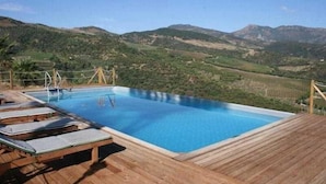 A rectangular swimming pool with a wooden deck and lounge chairs, surrounded by a fence, with a mountainous landscape in the background. - Tenuta Luogomarchese (Pollina)