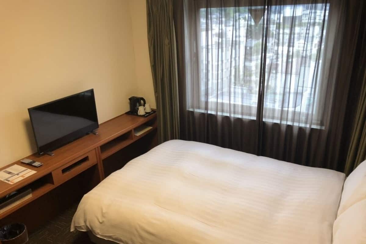A hotel room with a bed, a desk with a television, and a window with curtains.