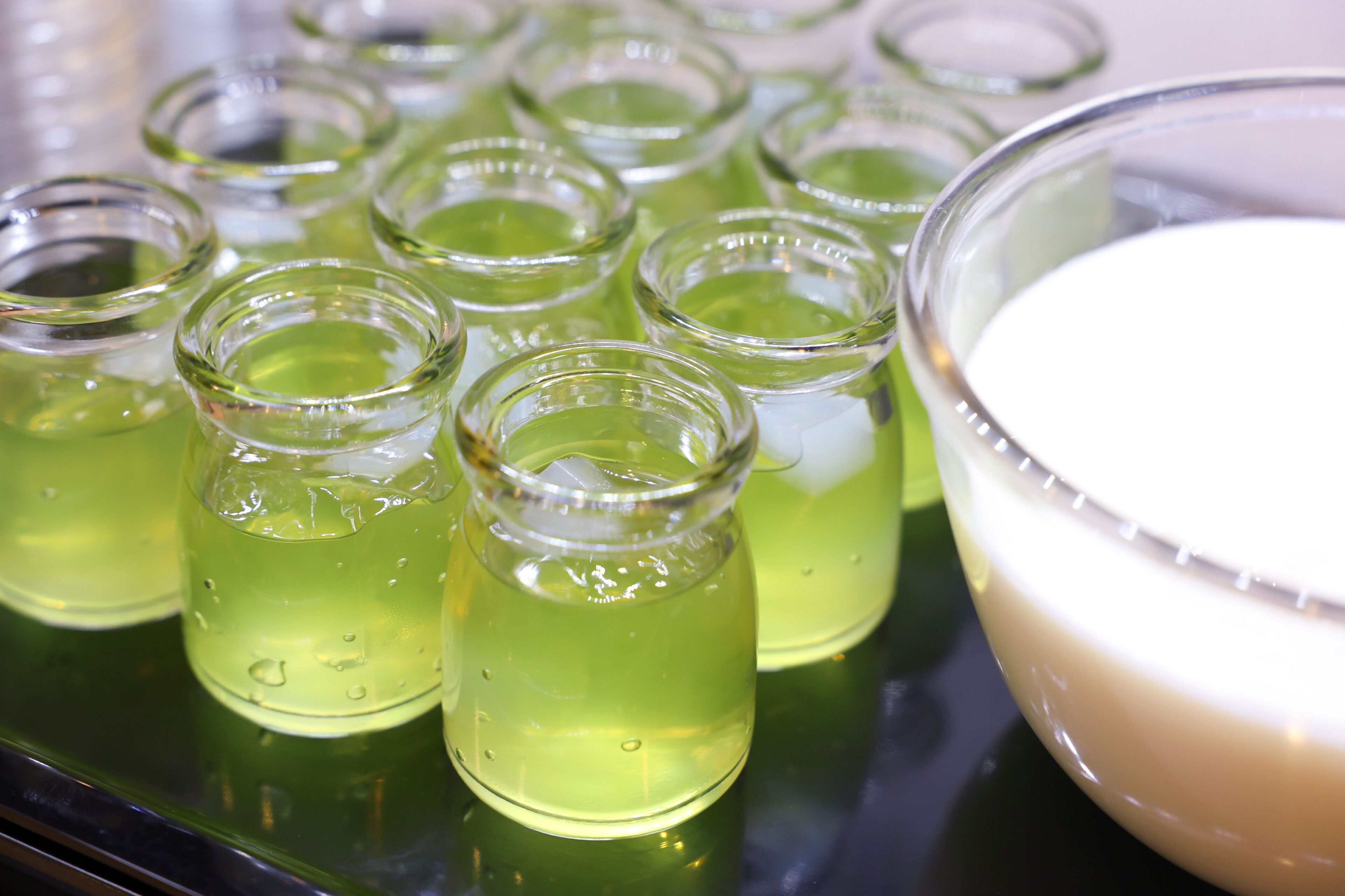 A tray with multiple small glass containers filled with a green liquid, likely a beverage, and a larger bowl with a white substance, possibly a food item, on a reflective surface.