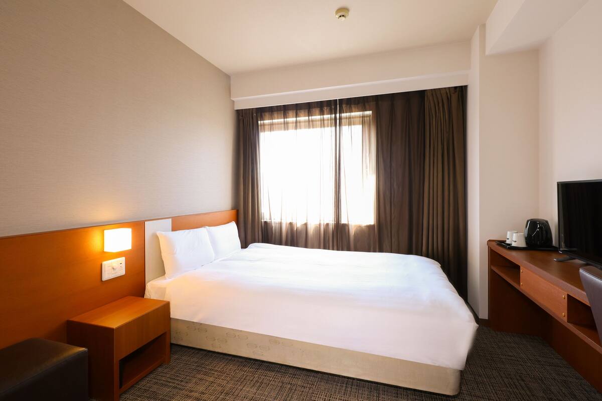 A hotel room with a large bed, a wooden nightstand, a desk with a chair, a television, and a window with curtains.