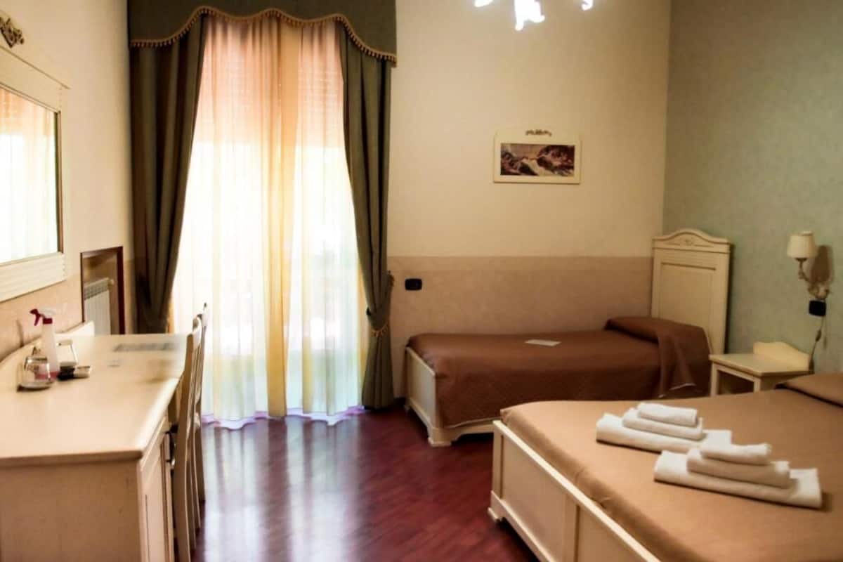 A hotel room with a bed, a bedside table, a chair, a desk with a lamp, and a window with curtains.