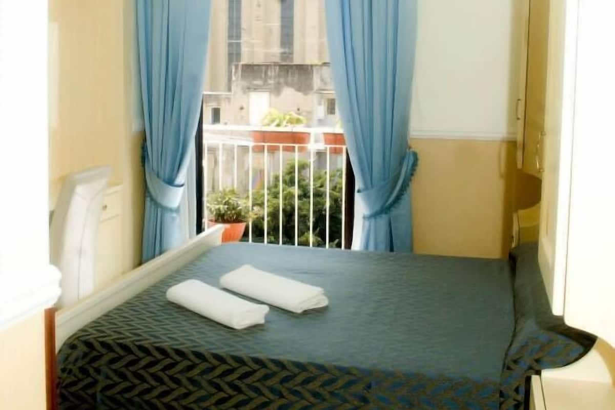 A small bedroom with a bed, a window with blue curtains, a chandelier, and a view of buildings outside.