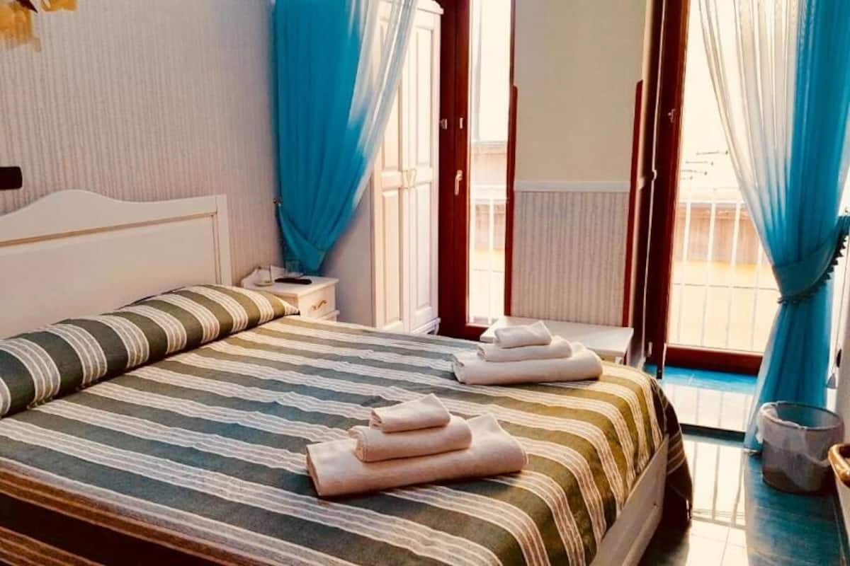 A hotel room with a bed, a nightstand, a window with curtains, and a view of the outside.