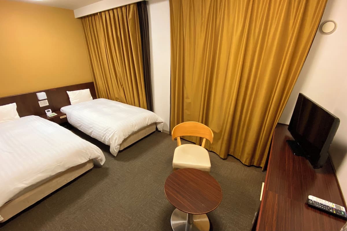 A hotel room with two beds, a wooden chair, a small round table, a TV, and curtains.