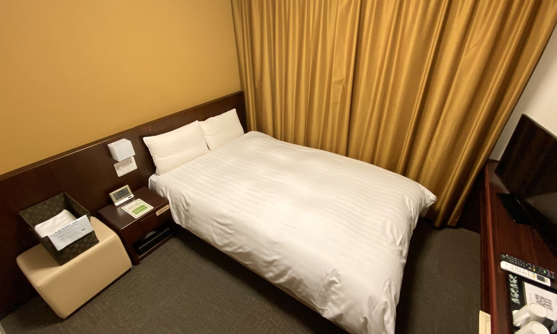 Habitación de hotel con cama, mesita de noche con un reloj, control remoto y una pequeña mesa con una caja de pañuelos y una nota.