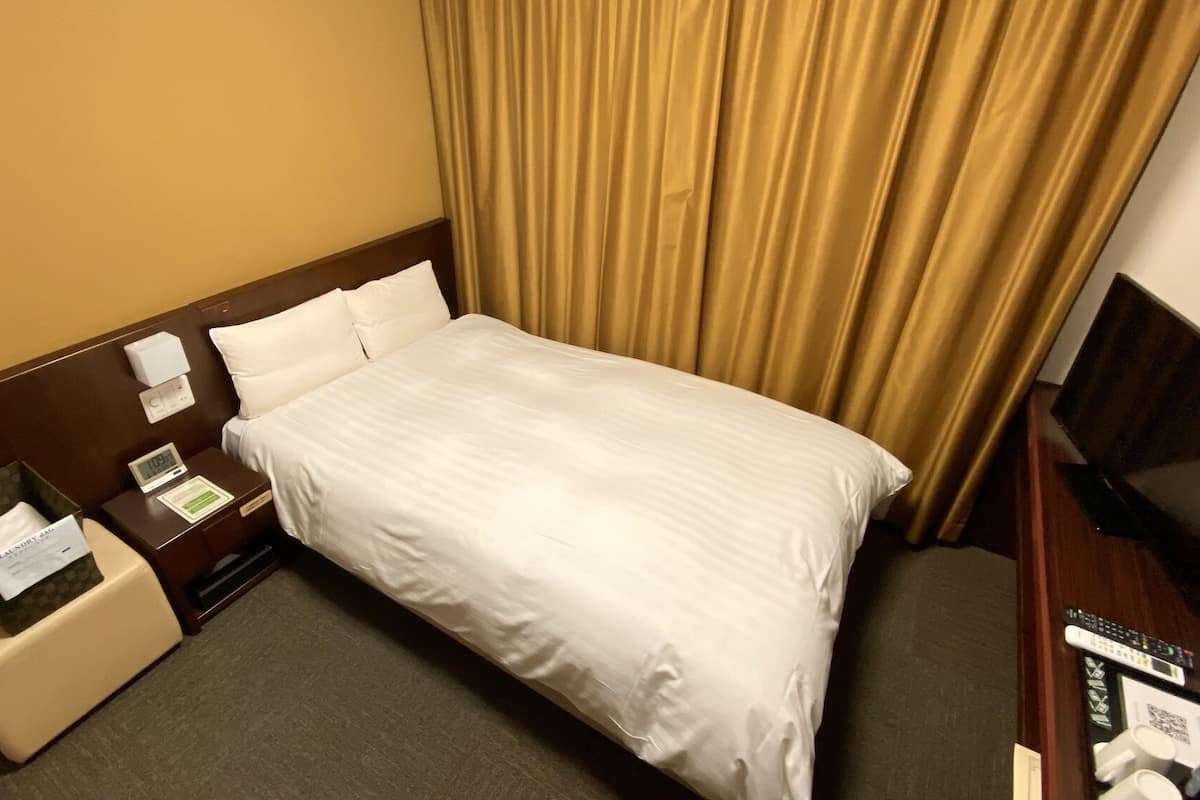A hotel room with a bed, a nightstand with a clock, a remote control, and a small table with a tissue box and a note.