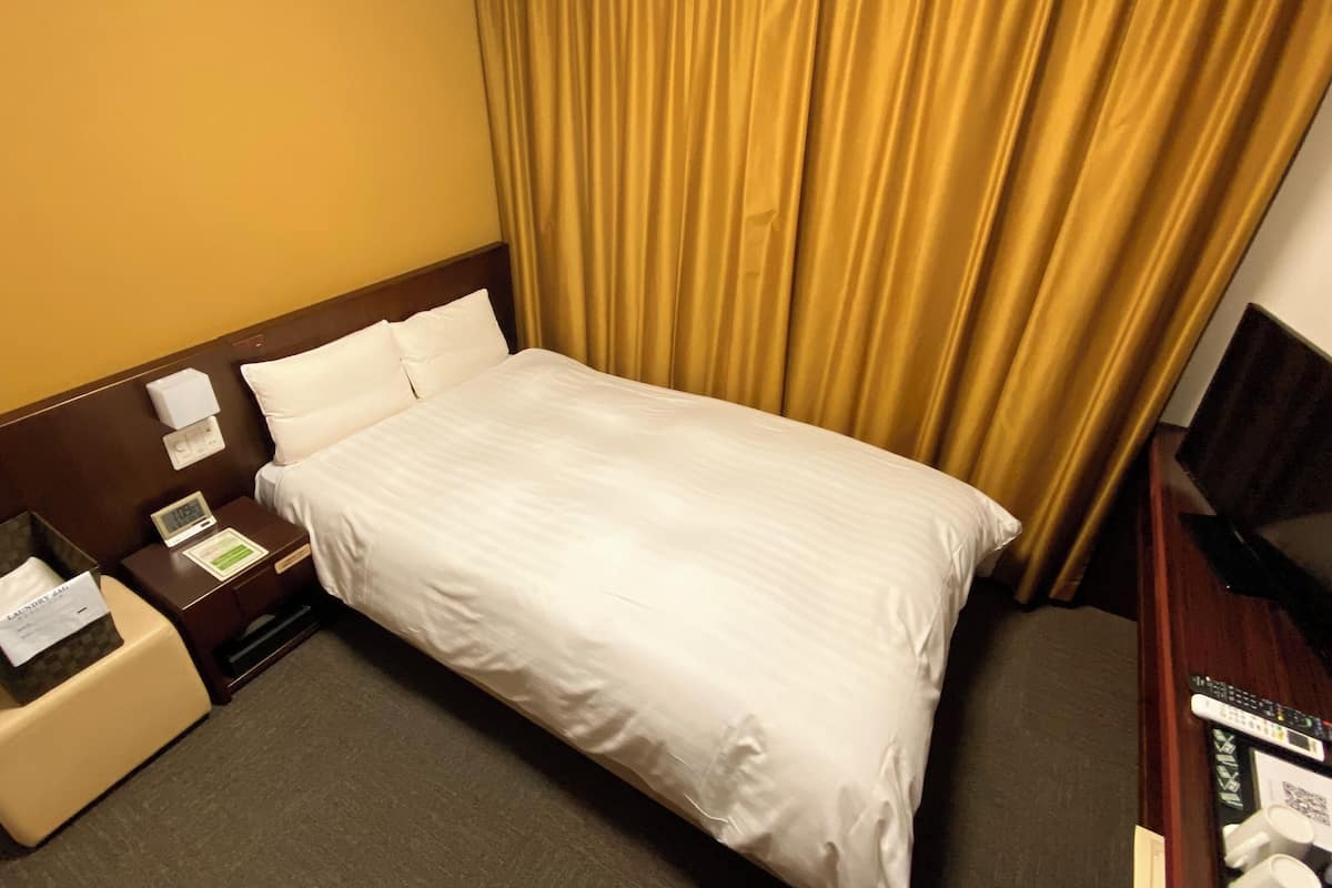 A hotel room with a bed, a nightstand with a clock, a small table with a basket, and a TV stand with a remote control.