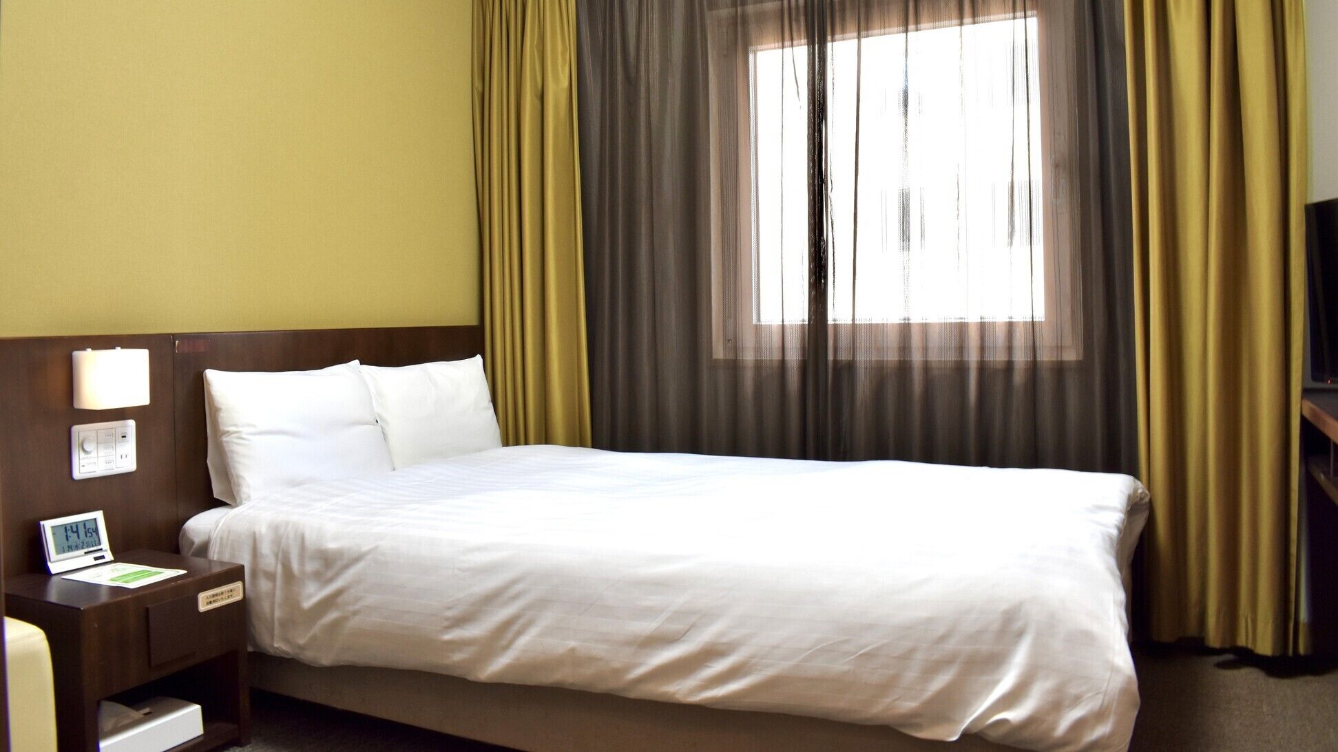 Una habitación de hotel con cama, mesita de noche con lámpara, reloj digital y ventana con cortinas.