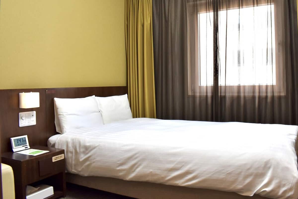 A hotel room with a bed, a nightstand with a lamp, a digital clock, and a window with curtains.