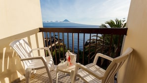 Ein Balkon mit zwei weißen Plastikstühlen, einem kleinen Tisch mit einer Flasche und einem Glas sowie Blick auf den Ozean und ein Gebirge.