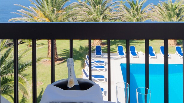Ein Tisch mit einer Champagnerflasche, zwei Gläsern und einer Schale Erdbeeren, mit Blick auf einen Pool und Palmen, im Hintergrund ein Berg.