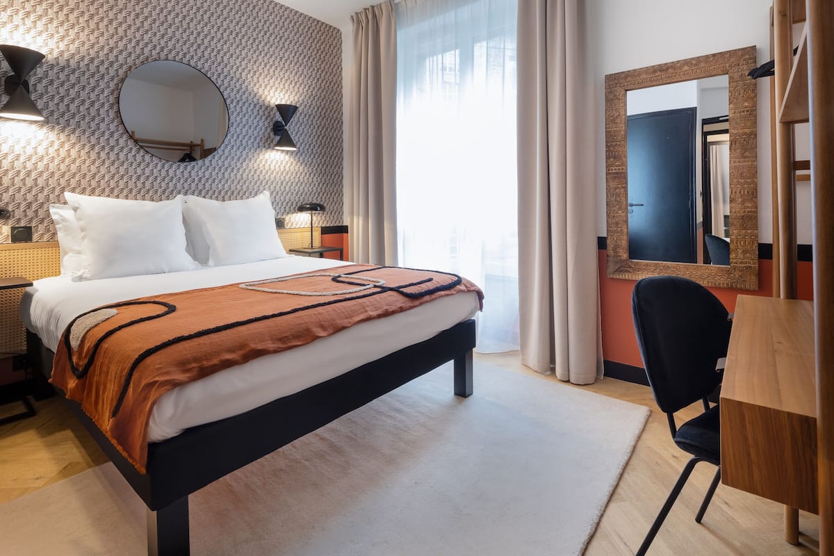 A hotel room with a large bed, a desk, a chair, a mirror, and a patterned wall.