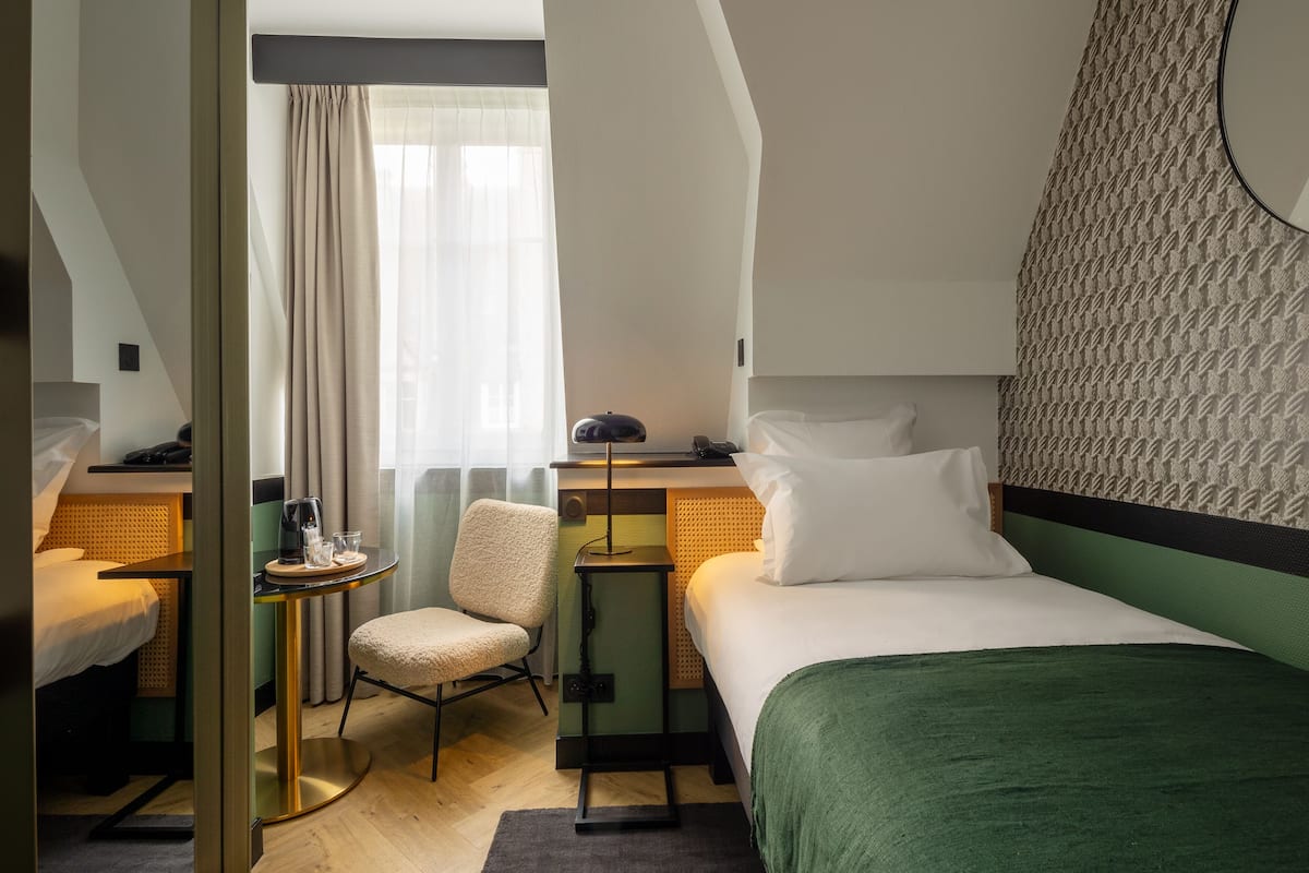 A hotel room with a bed, a desk with a chair, a small table with a lamp, and a window with curtains.
