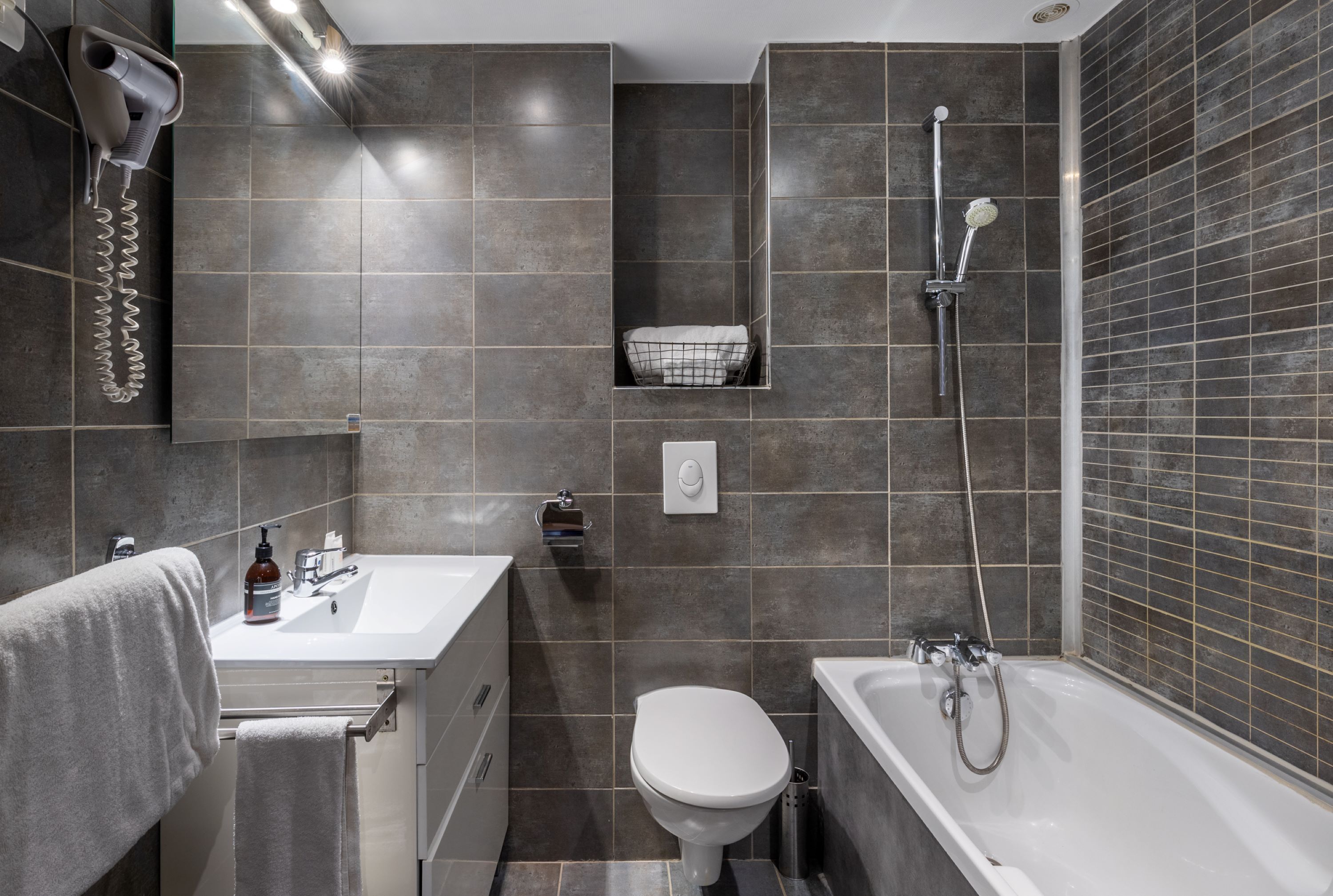 Un baño moderno con lavamanos blanco, inodoro, bañera y secador de pelo montado en la pared.