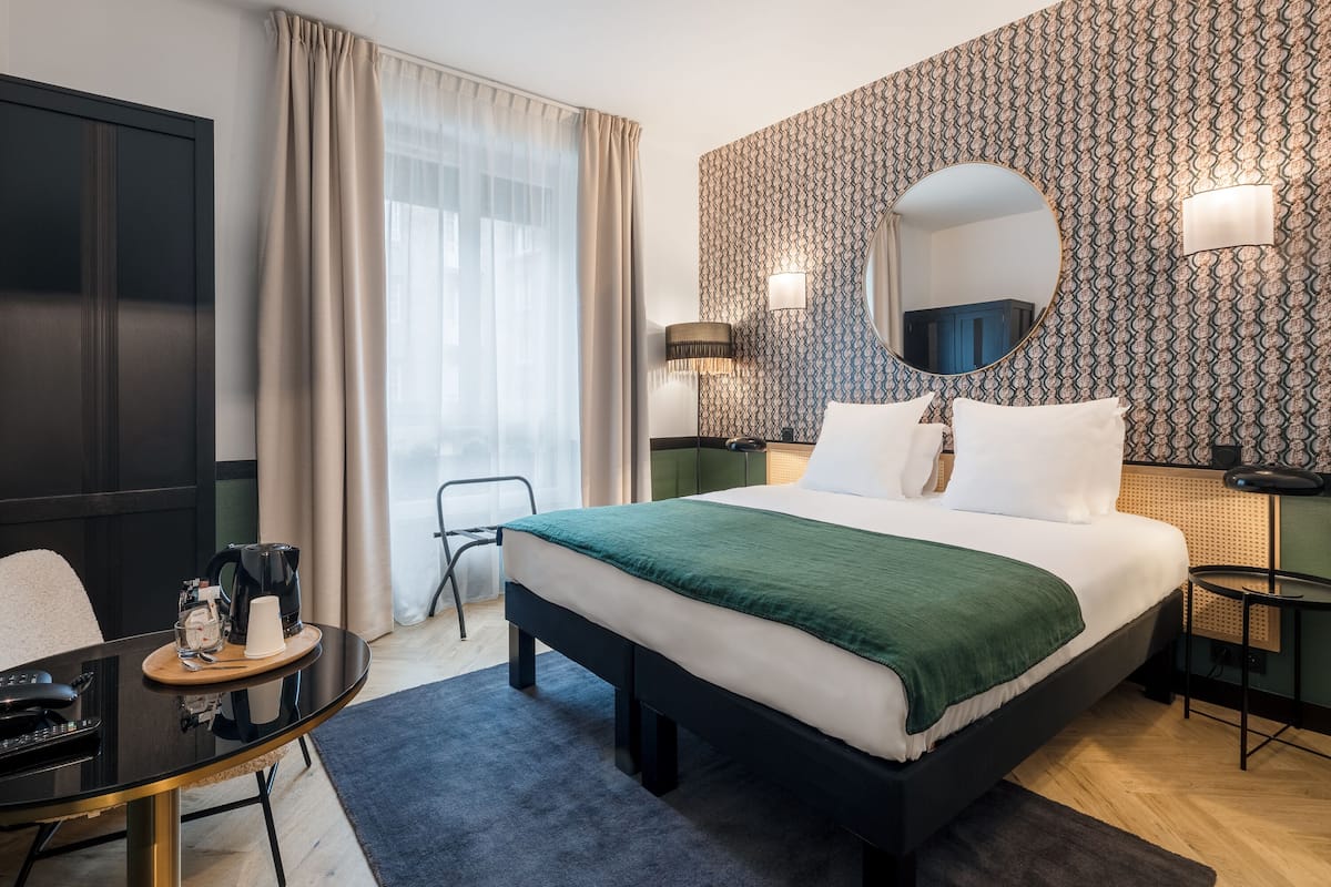 A modern hotel room with a large bed, a desk with a chair, a coffee maker, and a mirror on the wall.