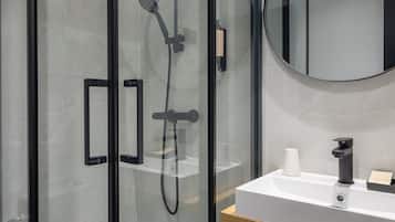 A modern bathroom with a glass-enclosed shower, a white sink, a round mirror, and neatly folded towels on a shelf.