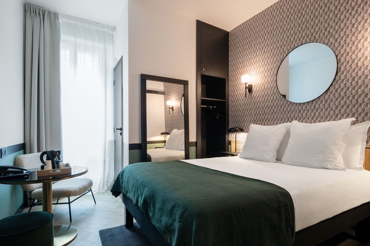 A hotel room with a bed, a round mirror, a small table with a chair, and a wardrobe.