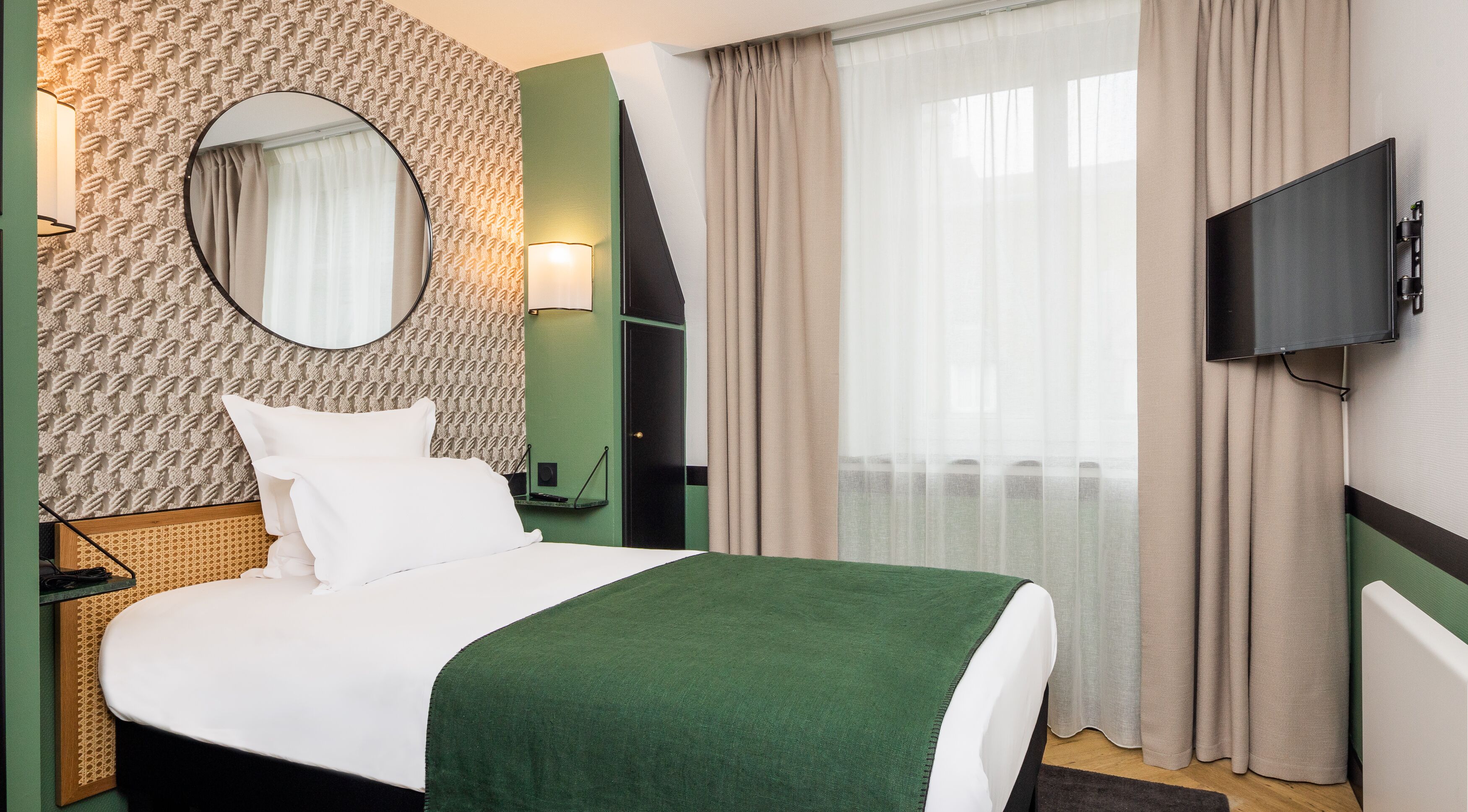 Une chambre d’hôtel avec un lit, un miroir rond, une télévision et une fenêtre avec des rideaux.
