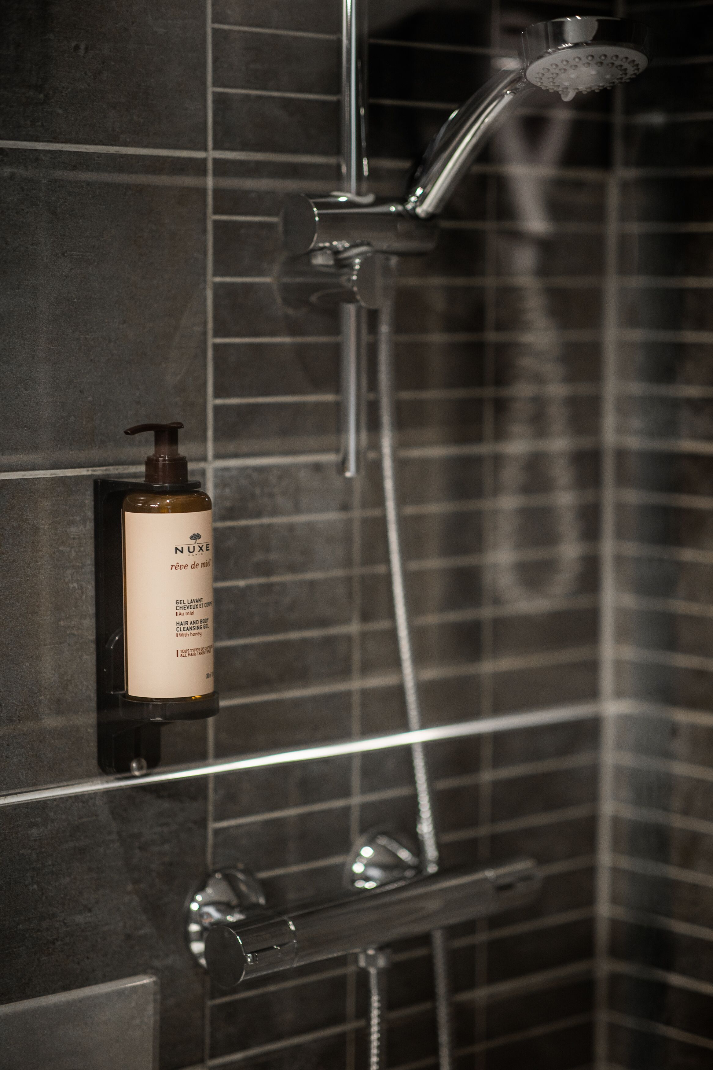A shower with a handheld showerhead and a bottle of Nuxe body wash on a tiled wall.