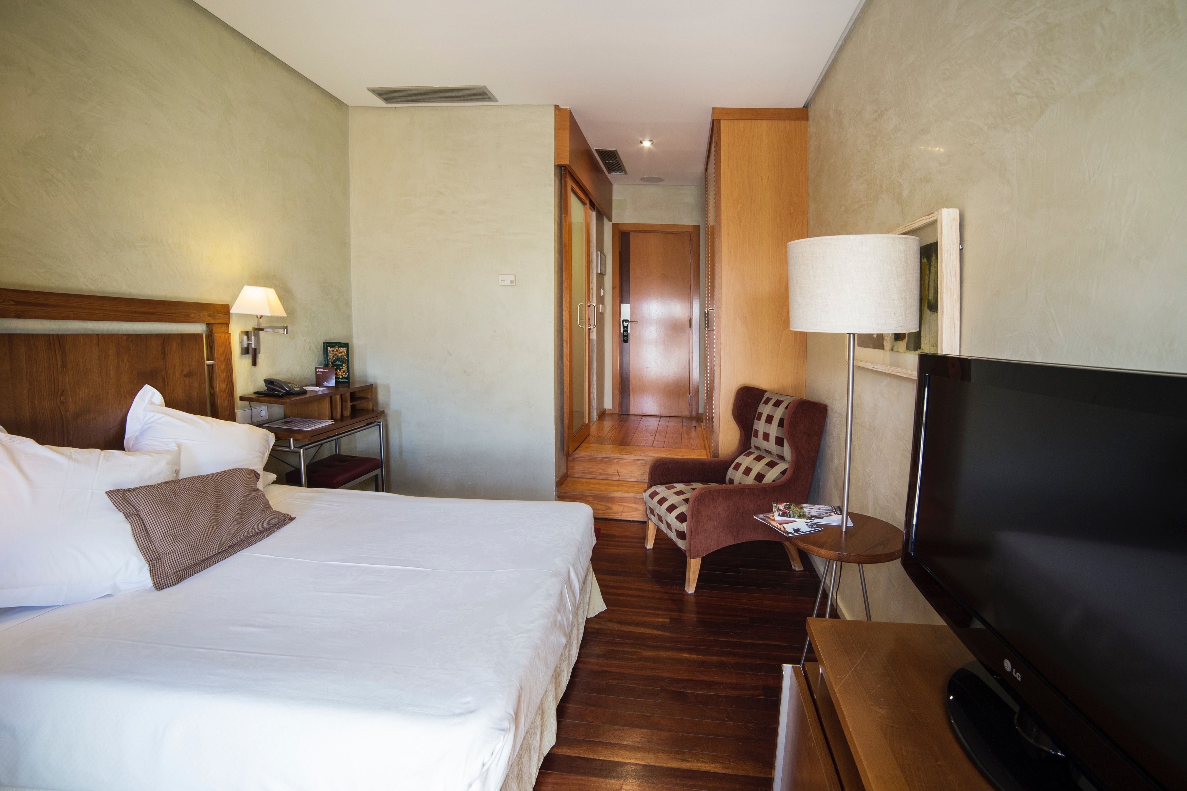 A hotel room with a bed, a television, a chair, and a desk with a lamp.