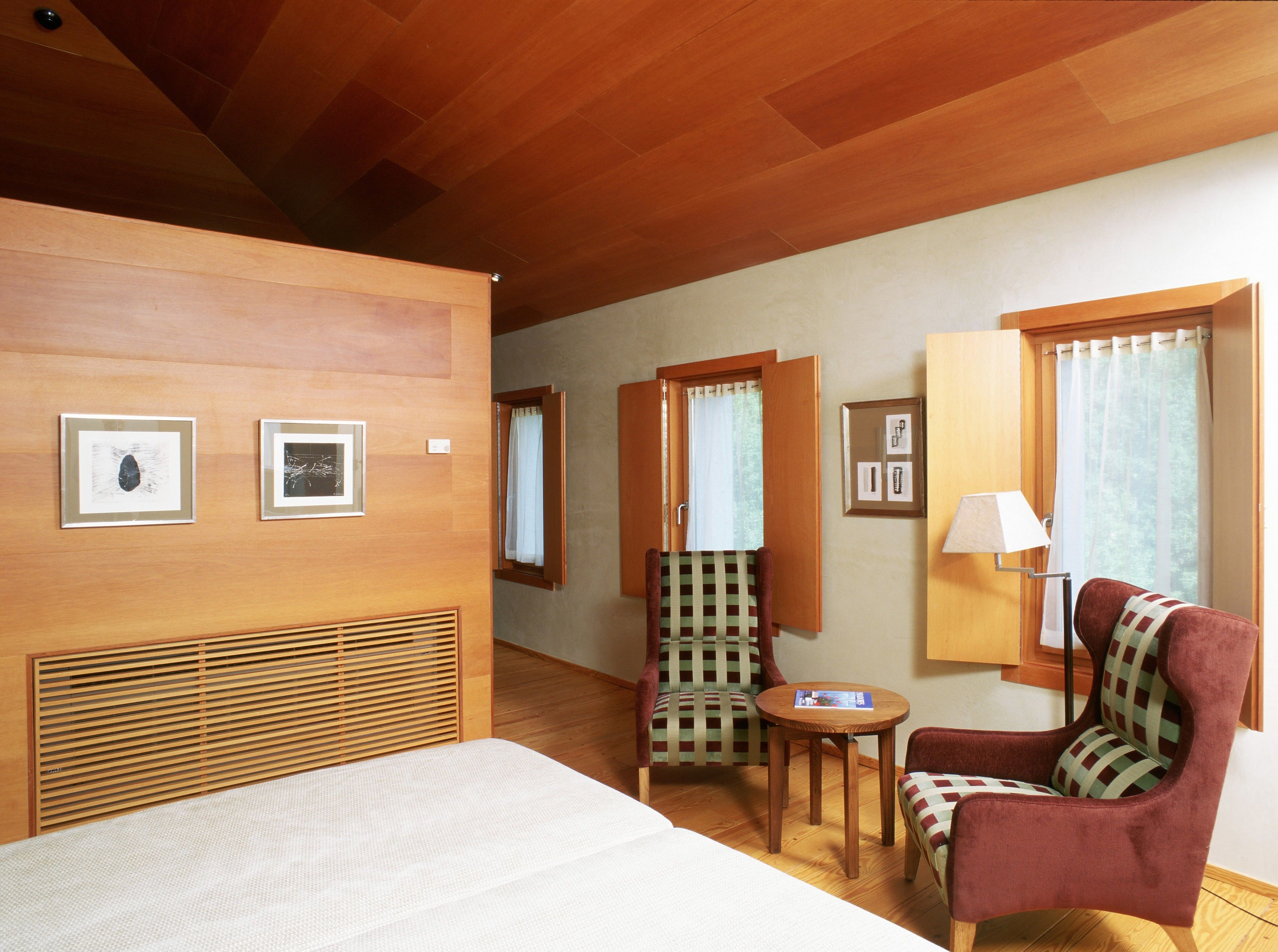 A hotel room with a bed, a chair, a small table, and a lamp. There are pictures on the wall and a window with curtains.