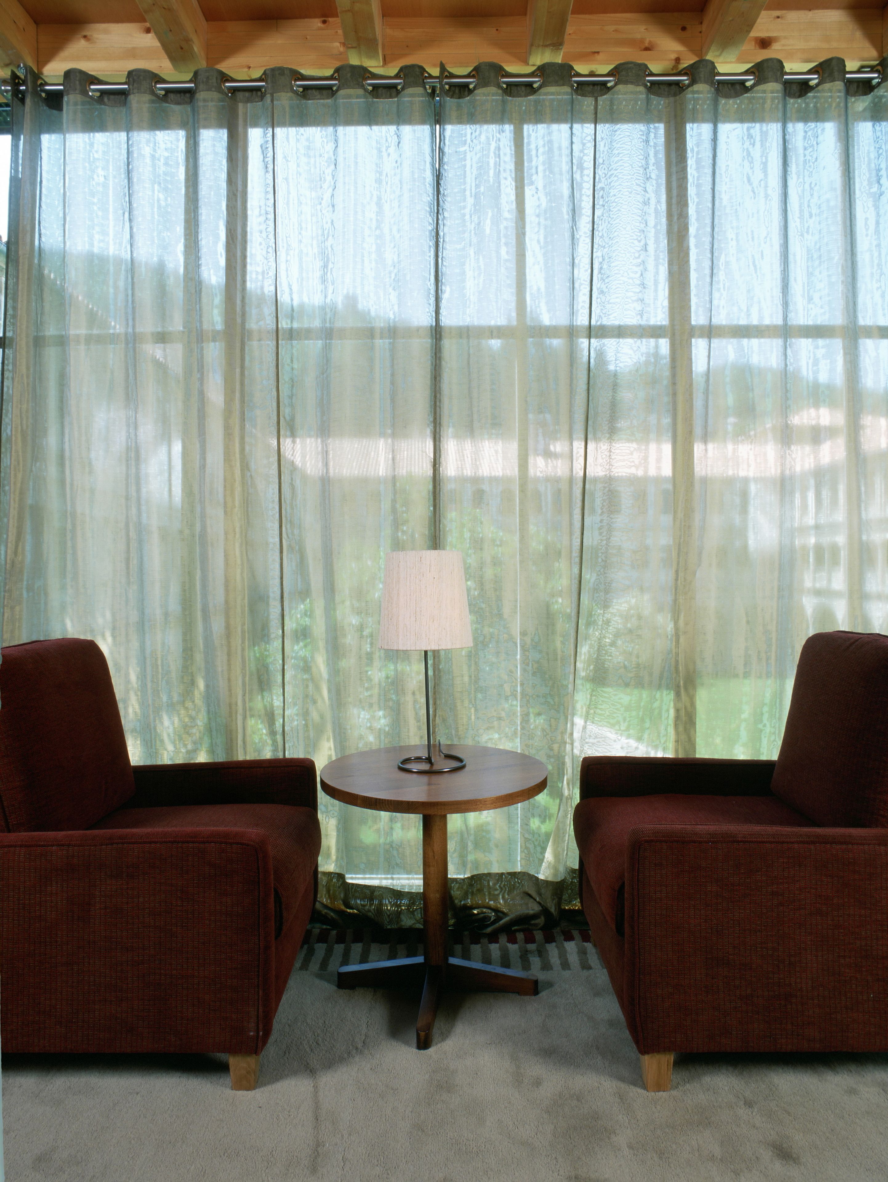A room with two armchairs, a small round table with a lamp, and sheer curtains.