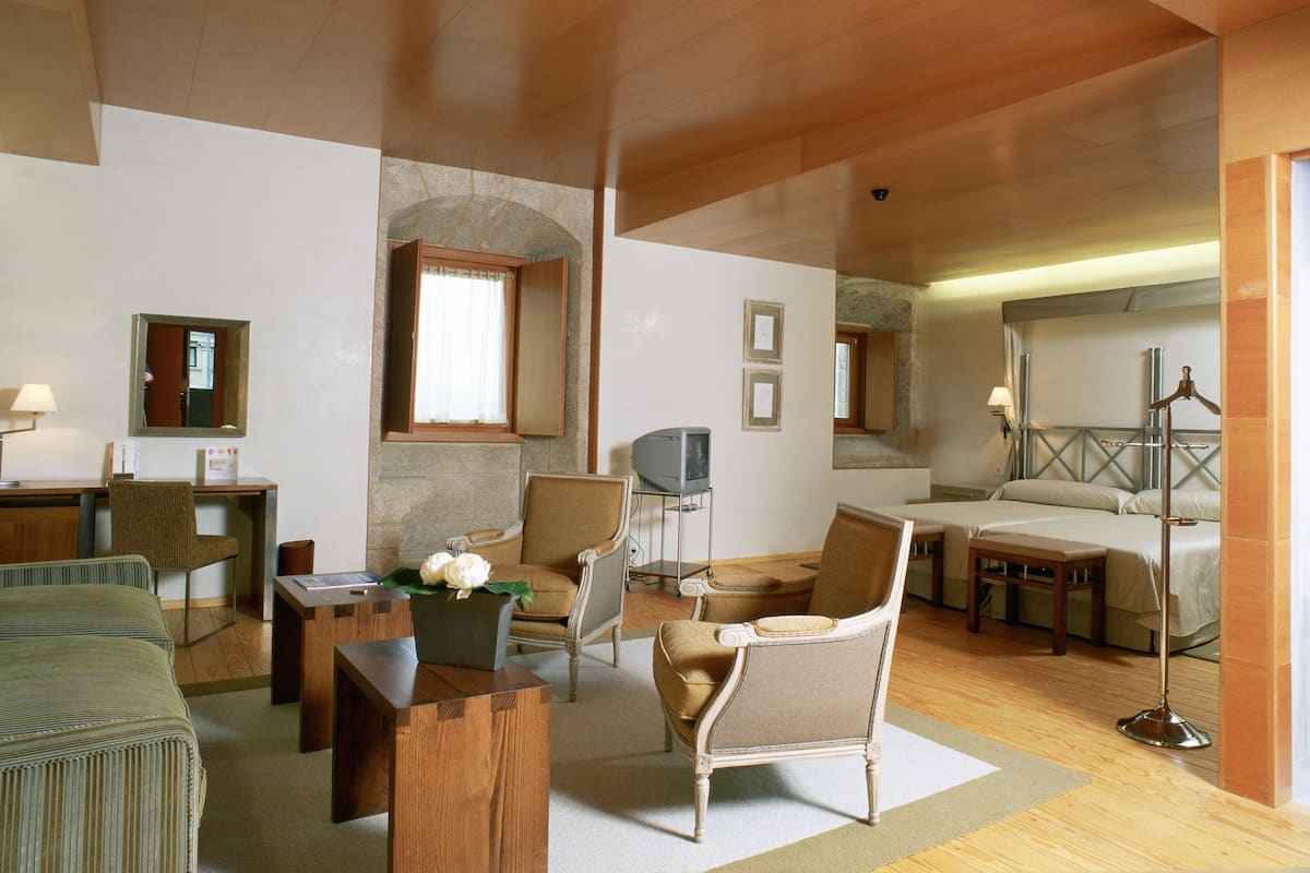 Ein Hotelzimmer mit Holzbalkendecke, einer Wand aus Naturstein, einem Fernseher, einem Schreibtisch mit Stuhl, einem Bett und einer Stehlampe.