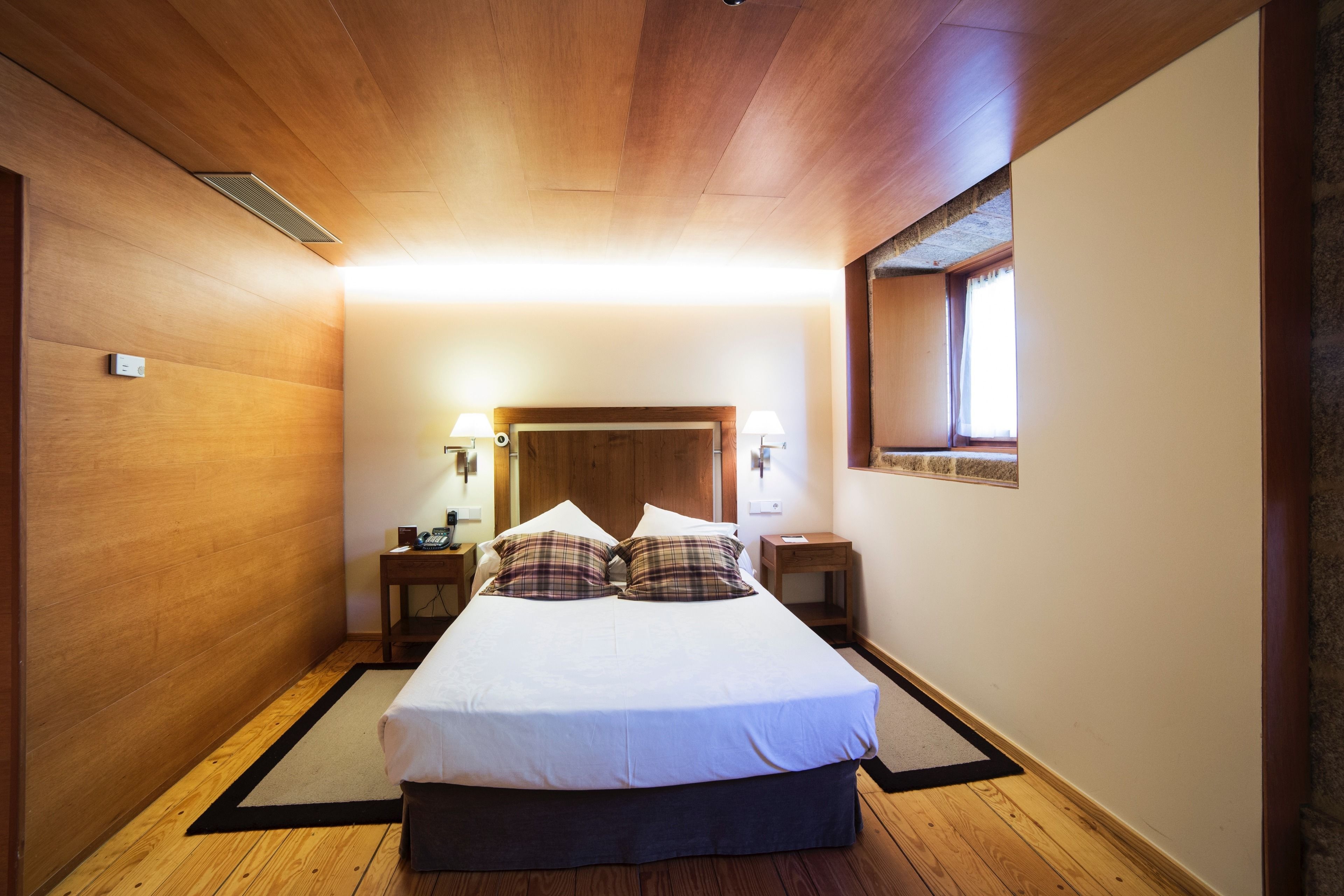 Une chambre avec un plafond en bois, un lit recouvert de linge de lit blanc, deux tables de chevet avec des lampes et une fenêtre donnant sur un beau paysage.
